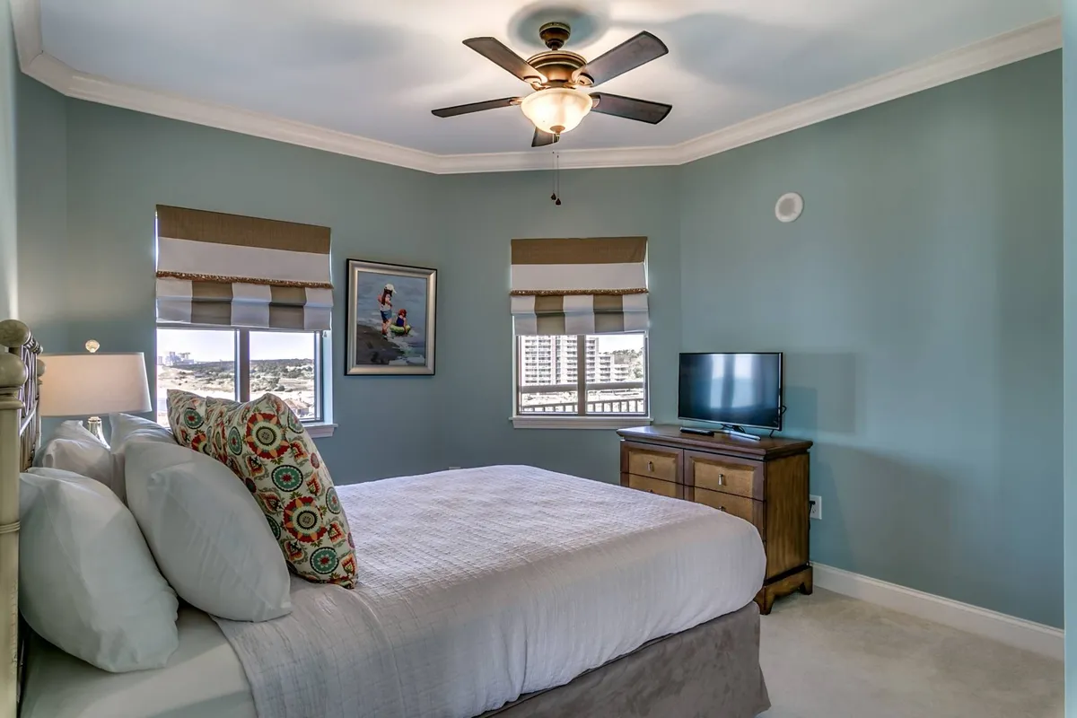 A cozy queen bedroom featuring a bed with decorative pillows, a TV on a wooden dresser, and windows with shades letting in soft natural light. Wall art adds a touch of personality to the peaceful space.