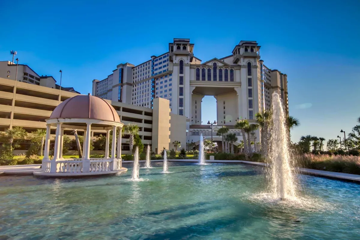 A grand resort entrance framed by an ornate gazebo and fountains, surrounded by manicured landscaping. Guests can be seen enjoying the tranquil water features and the elegant atmosphere, making for a welcoming arrival experience.