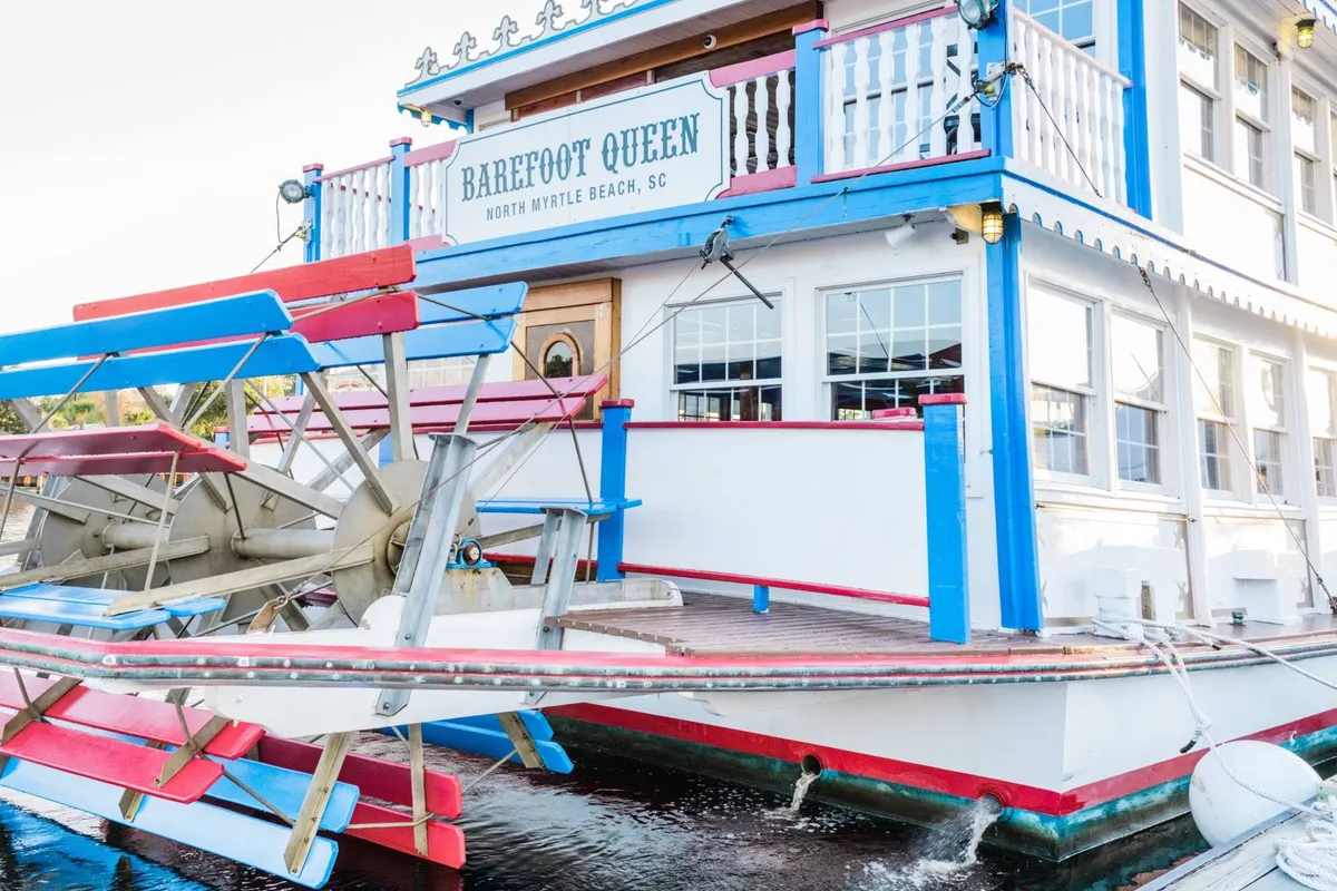 A brightly colored paddle wheel riverboat stands docked alongside lively signage and a vibrant building façade. The setting sun hints at upcoming evening cruises, while the boat's decks promise scenic tours, dining, and entertainment on the water.