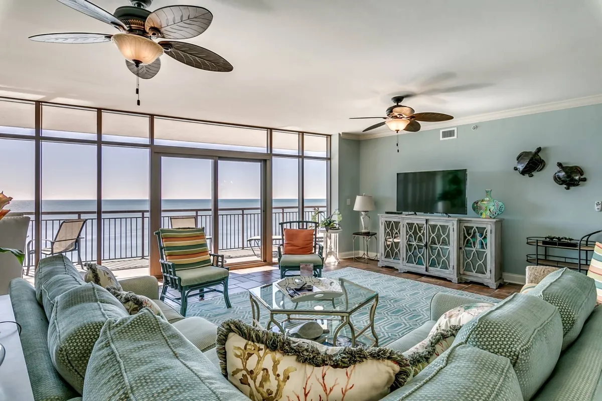 A bright and spacious living area featuring a comfortable sectional sofa, colorful accent chairs, and a sleek coffee table. The room is anchored by large floor-to-ceiling windows that frame sweeping ocean views, seamlessly connecting the interior with the natural beauty outside. The mood is casual and cheerful, perfect for relaxation or entertaining guests.