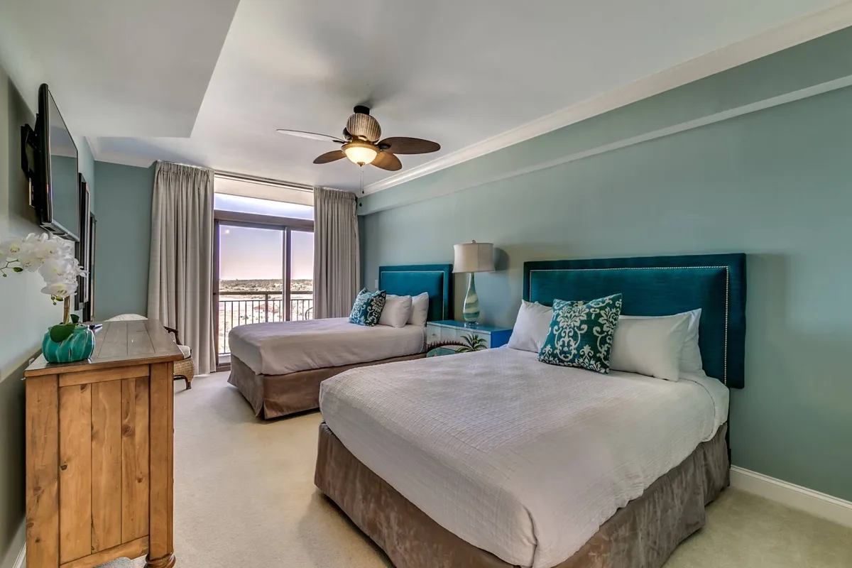 A bright bedroom featuring two queen beds with stylish teal headboards, a wooden dresser, a large window draped with curtains, and a ceiling fan overhead. The space exudes a calm, restful vibe perfect for families or groups.