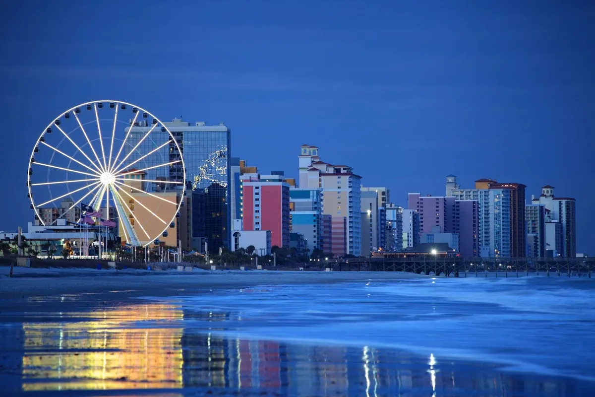 A vibrant, festive beachfront scene featuring a towering Ferris wheel, colorful buildings, and the sun setting over the ocean, reflected in the water below. The area is alive with the energy of seaside attractions and nighttime strolls.