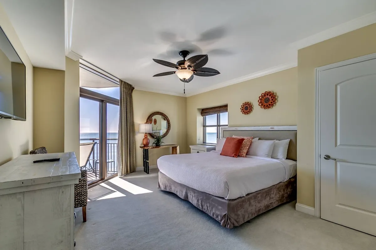 A spacious king-sized bed dominates the room, flanked by large windows with serene ocean views. Decorative wall art and a ceiling fan contribute to a relaxing atmosphere, while a TV sits opposite the bed, ideal for a cozy night in.
