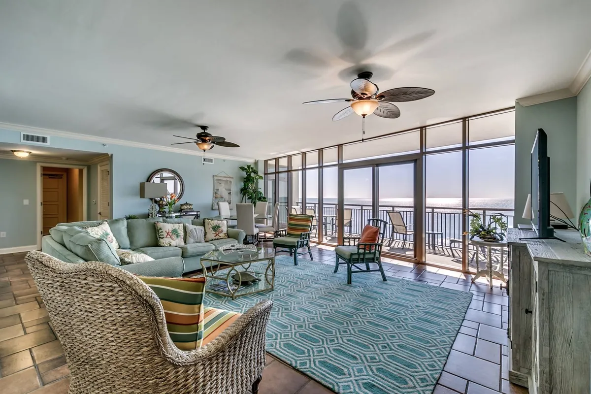 The image showcases a bright, spacious living area with large windows offering stunning views, a welcoming seating area perfect for gathering, lush decorative plants, and a vibrant area rug that adds a touch of coastal charm.