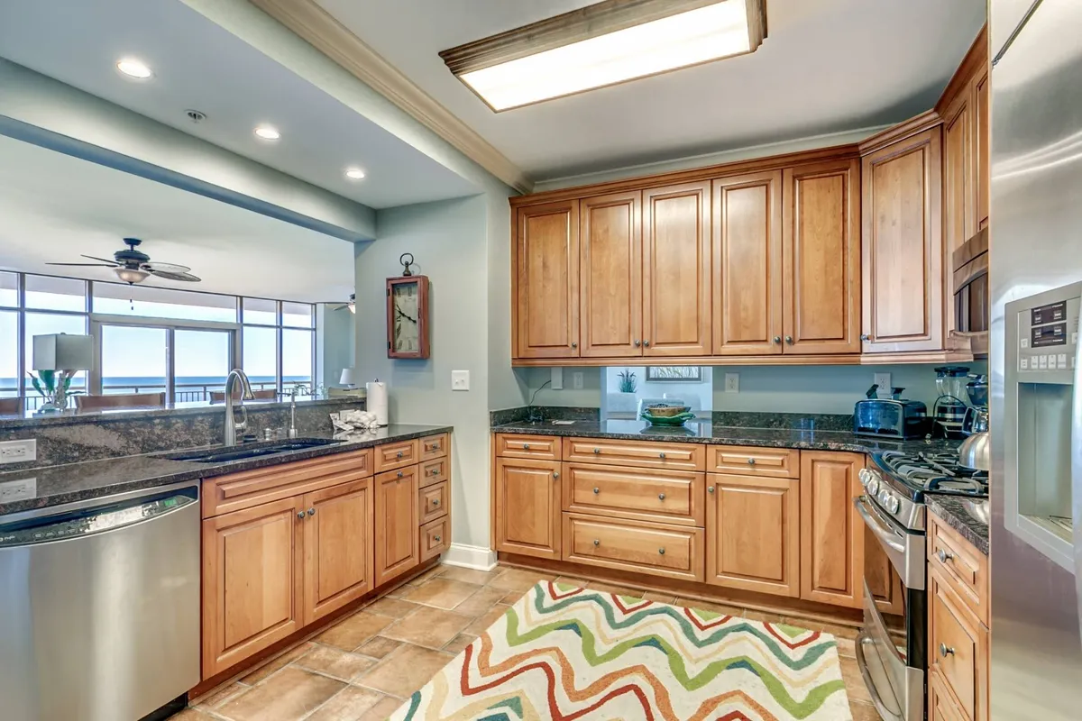 A spacious modern kitchen featuring granite countertops, wooden cabinets, a colorful area rug, and a full suite of stainless steel appliances. The layout is open, inviting social interaction while cooking. The kitchen is set up for both casual and celebratory meals, with hints of cookware and small appliances visible.
