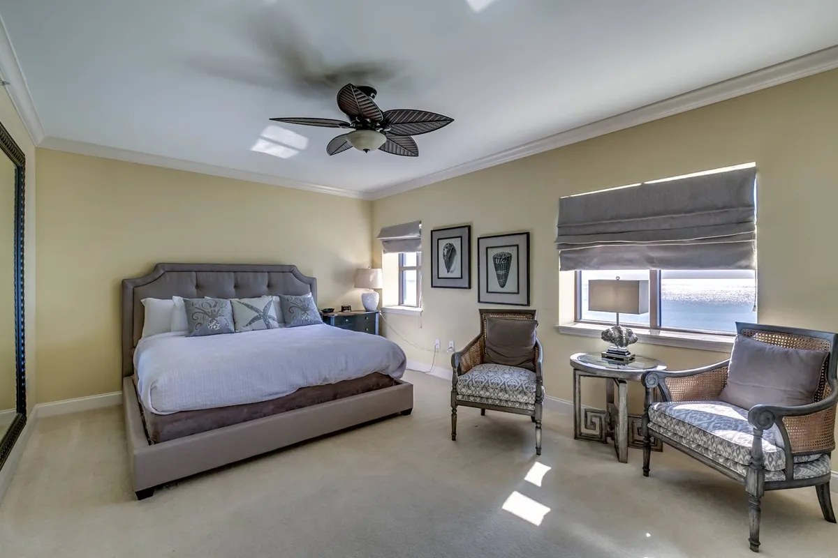 This image shows a tranquil bedroom space with a comfortable bed, armchairs, a lamp, framed art, and a mirror. Morning light streams through the windows, hinting at views of the sunrise and creating a cozy, inviting atmosphere for relaxation throughout the year.
