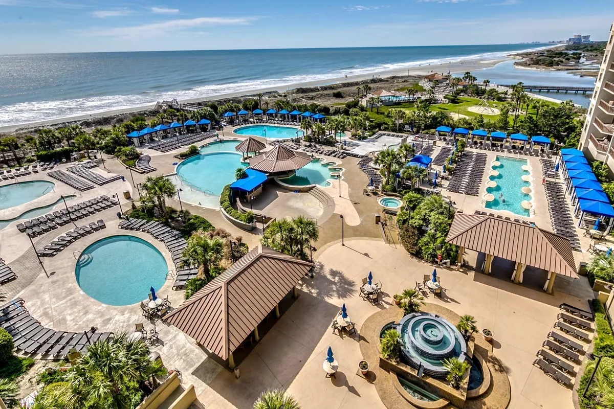 This exterior image showcases the expansive swimming pools surrounded by landscaped gardens and palm trees, with seating areas for lounging and an ocean view in the background. The scene captures people swimming, sunbathing, socializing, and relaxing by the pool, evoking a festive and leisurely resort atmosphere.