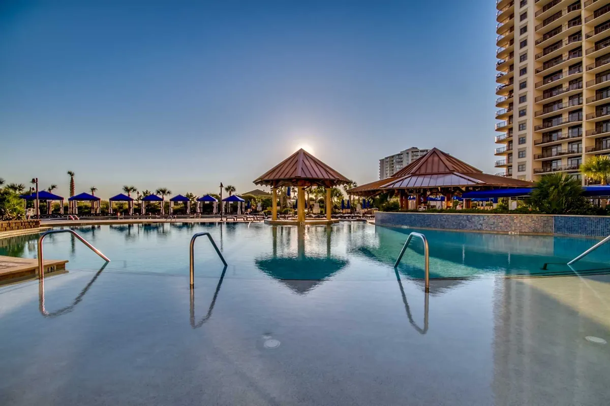 A luxurious resort swimming pool surrounded by swaying palm trees and tiki-style cabanas, with the scene softly illuminated by the setting sun. Guests are seen relaxing by the pool and socializing, creating an inviting atmosphere for families and adults alike.
