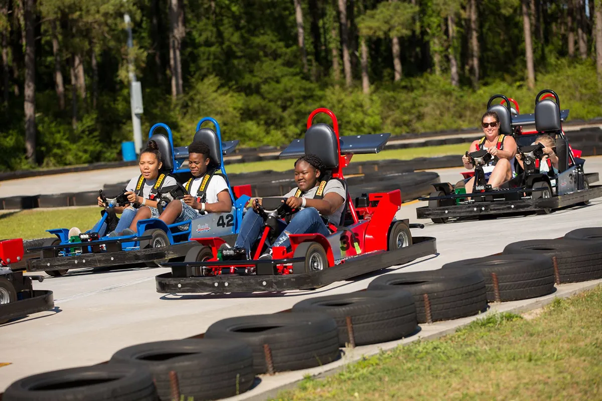 Participants of all ages race around a dynamic go-kart track, the excitement palpable in their expressions. The scene is energetic, filled with the blur of speeding karts, laughter, and competition, set against the backdrop of a well-designed family attraction.